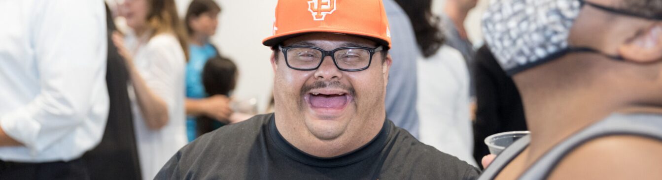 Close up of a man wearing an orange hat and glasses captured mid laugh.